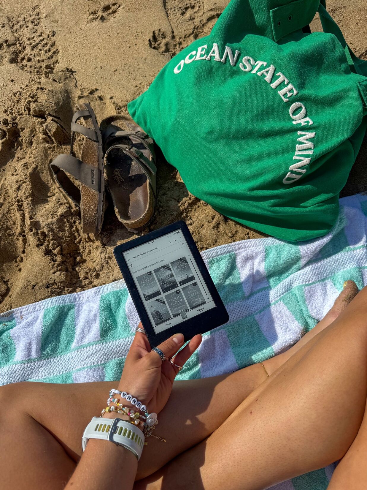 legs of girl sitting on a towel at the beach reading on kindl and green bag